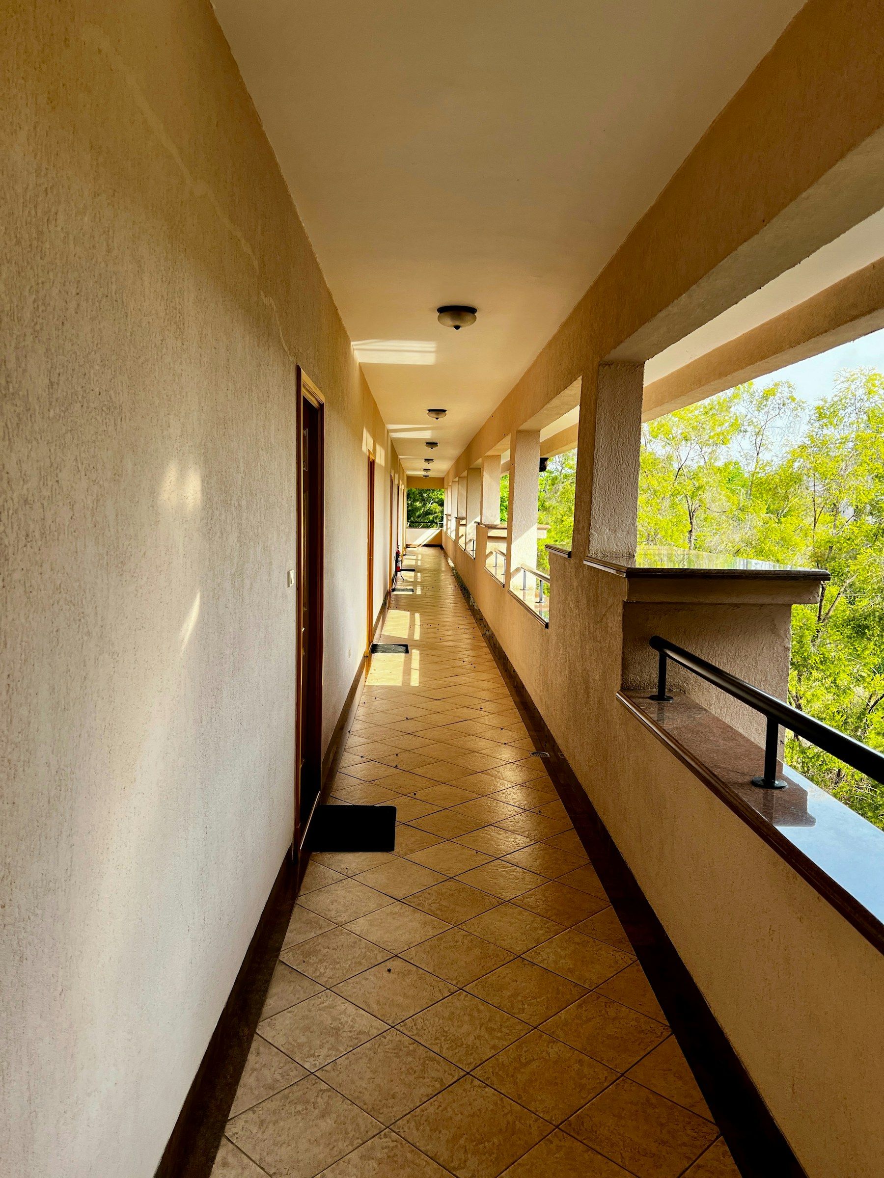 An open hotel corridor with room doors on one side and trees on the other